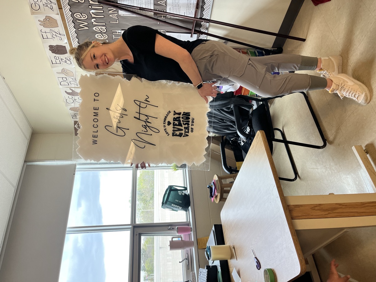 Danielle in her classroom holding a hand-made acrylic sign from her Mrs. P Creations sign business